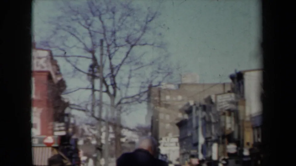 1959 New York street scene showing bare trees, mixed building facades, pedestrians in coats, traffic, and home-movie frame borders.