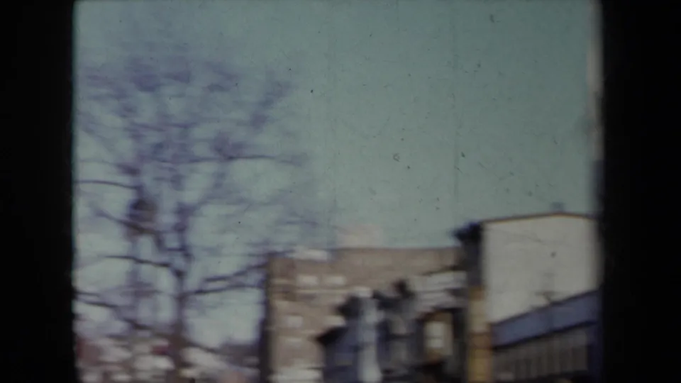 Soft 8mm frame of a 1959 New York streetscape with bare tree branches, low buildings, pale sky, and blurred urban frontage.