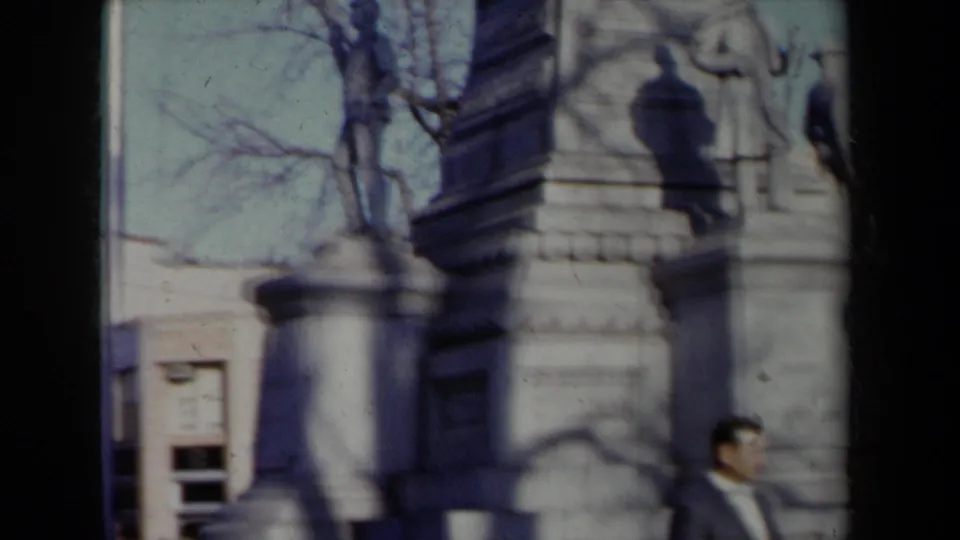 1959 New York frame with a large stone monument or memorial base, surrounding buildings, bare trees, and a pedestrian at the lower edge.
