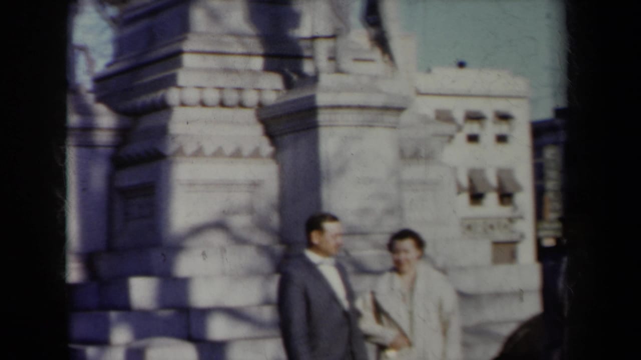 In 1959 New York home-movie footage, a suited man and light-coated woman stand near a stone monument with carved figures.