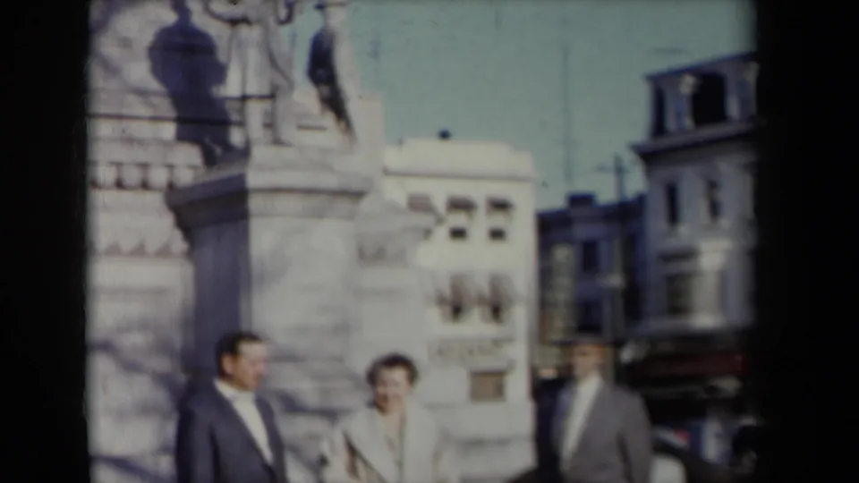 A suited man, a woman in a light coat, and another man pass a large stone monument on a New York street.