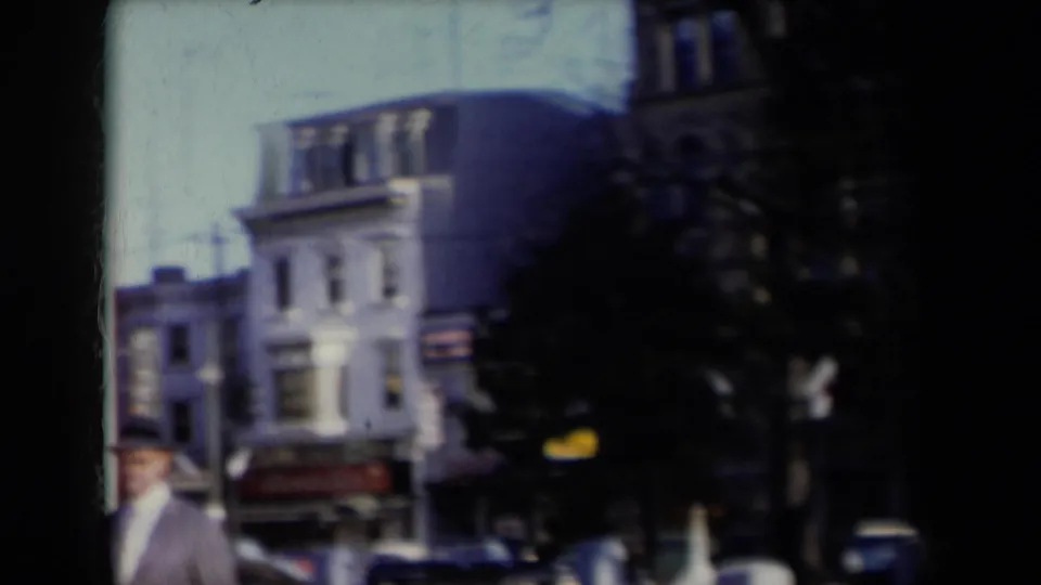 The home-movie camera pans toward nearby New York buildings, storefront awnings, parked cars, and a partially visible suited man.