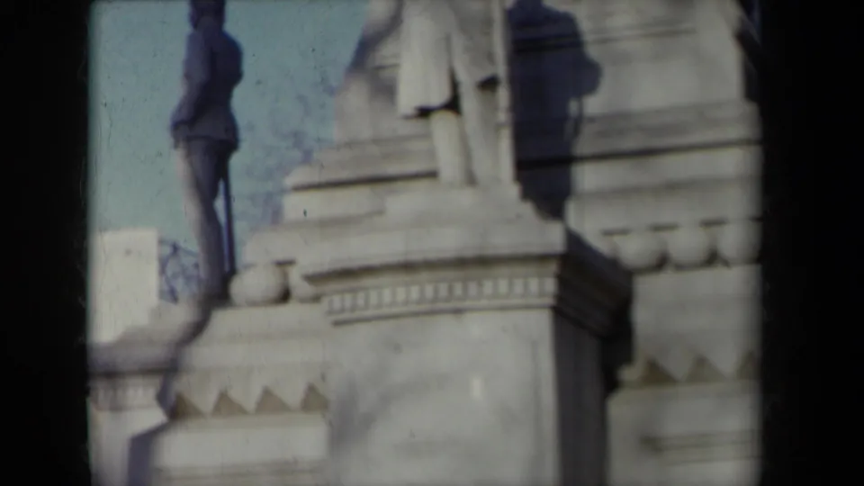 A suited man appears beside the monument base while a carved stone figure and architectural detailing fill the frame.