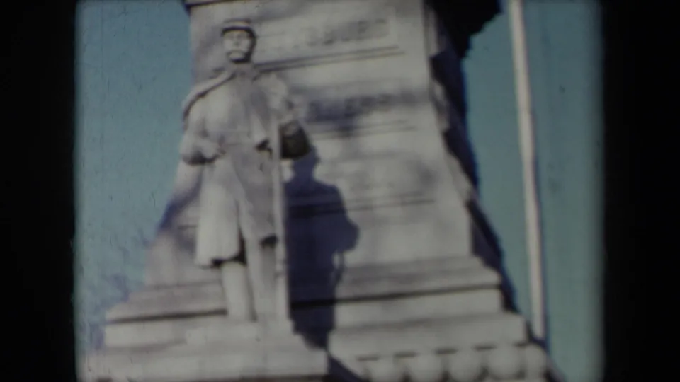 1959 New York 8mm frame showing a uniformed stone soldier figure beside a monument base, flagpole, carved lettering, and blue sky.