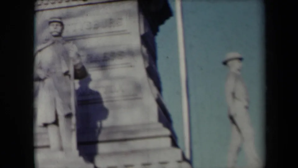 1959 New York 8mm frame showing the monument with a central soldier statue, a second side figure, carved base, flagpole, and blue sky.