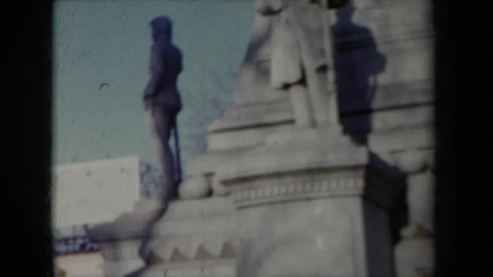 8mm home-movie frame from New York in 1959 showing a darker statue beside a pale tiered monument base under blue sky.