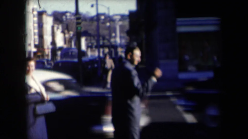 1959 New York street frame with a suited pedestrian near a crosswalk, older cars, storefront windows, and street poles.