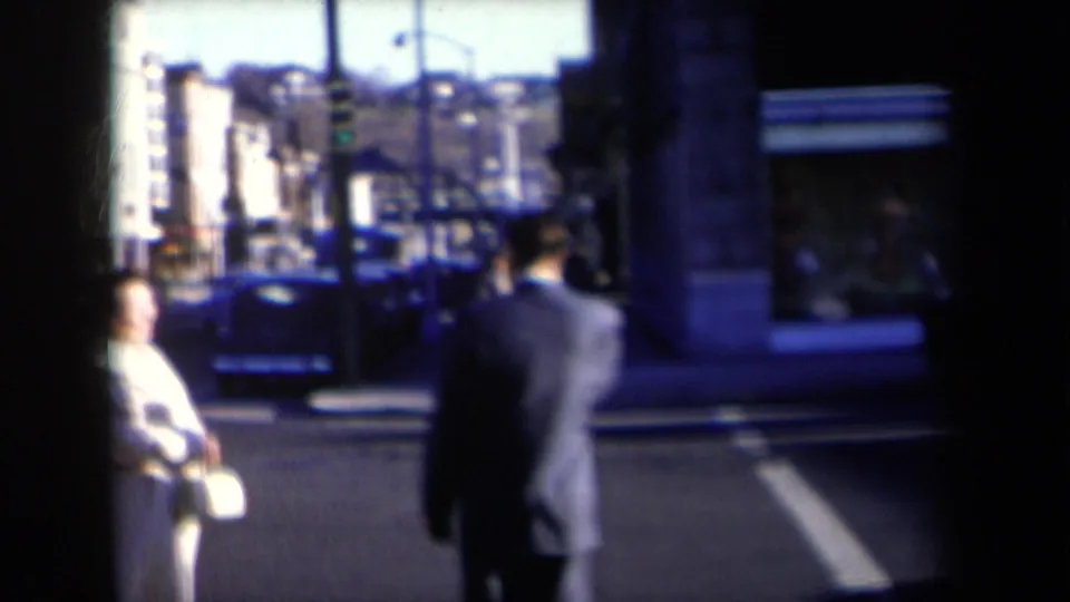 1959 New York street view showing pedestrians at a crosswalk, including a woman in a light coat and a suited man seen from behind.