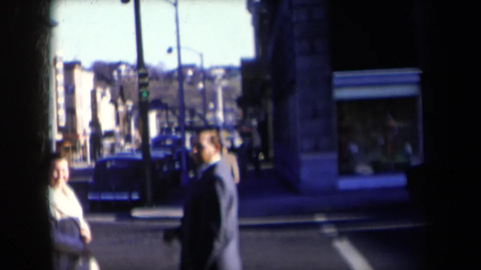 1959 New York frame with a suited man and a woman in a light coat near a crosswalk, with classic cars and storefronts behind them.