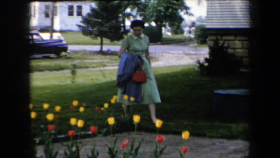 In 1959 New York home-movie footage, a woman in a light green dress carries a blue coat and red handbag beside tulips in a residential yard.
