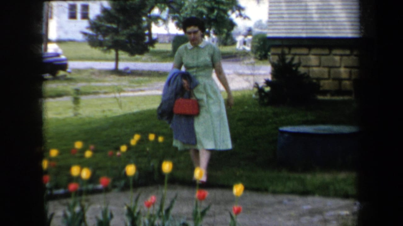 The woman in a light green dress pauses beside red and yellow tulips, with suburban houses, lawn, and a dark car visible in the background.