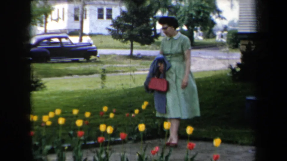 A woman in a green 1950s-style dress looks down at the tulip bed while standing on a garden path in the New York residential scene.