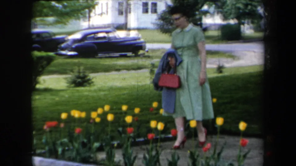 The woman remains beside the red and yellow tulips, holding a blue coat and red handbag with a parked dark sedan and homes behind her.