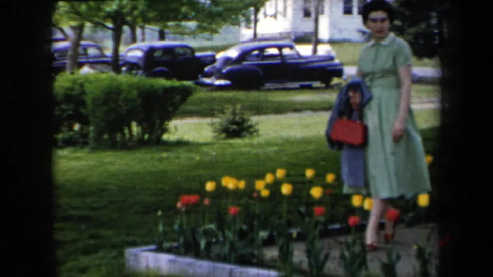 In 1959 New York home-movie footage, a woman in a green dress stands near tulips on a lawn with dark mid-century cars along the street.