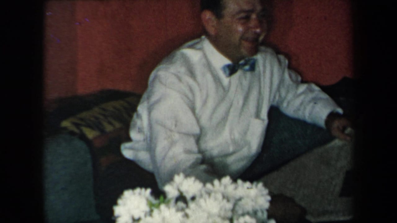 Man in a white shirt and bow tie sits on a couch beside a vase of white flowers.