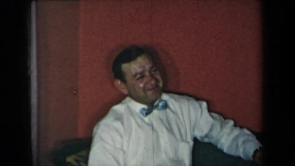 A seated man in a white shirt and bow tie smiles in a mid-century private room, framed by soft 8mm color and dark edge shadows.