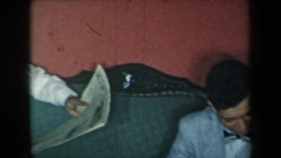 Inside a private room, a man leans near a couch while another partially visible person holds a newspaper, captured in dim 8mm footage.