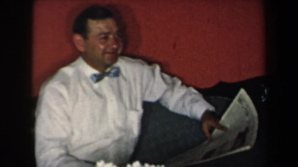 In 1959 New York home-movie footage, an adult man sits by a red wall holding a newspaper, with white flowers in the foreground.