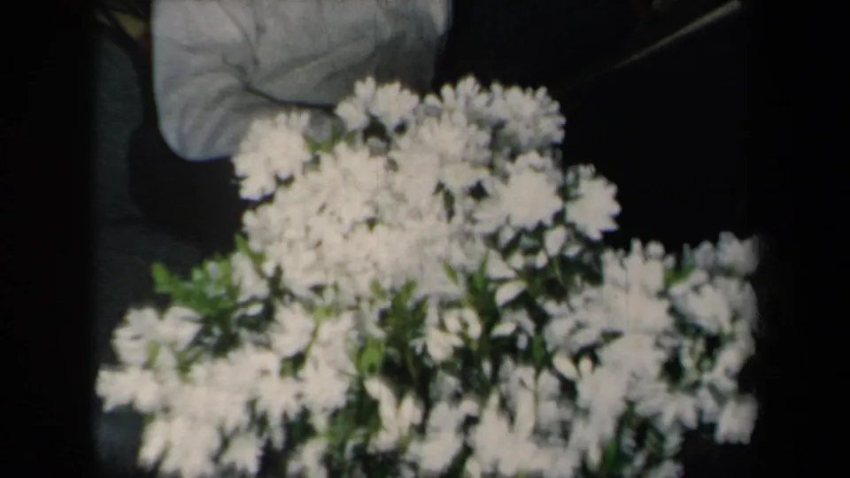 A dense white floral arrangement fills the lower frame, with a partly visible adult seated behind it in soft 8mm focus.