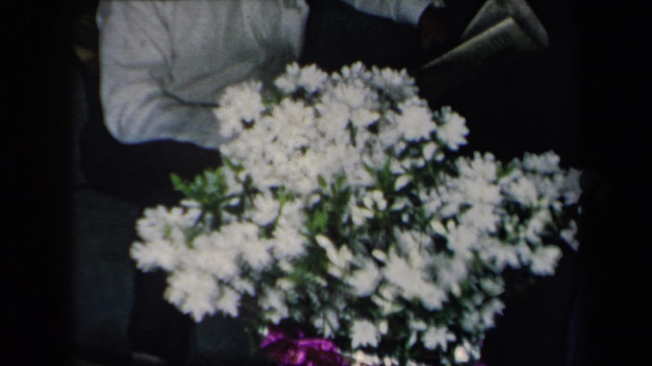 1959 New York 8mm frame showing white flowers in a shiny purple vase, with a partially visible person nearby.