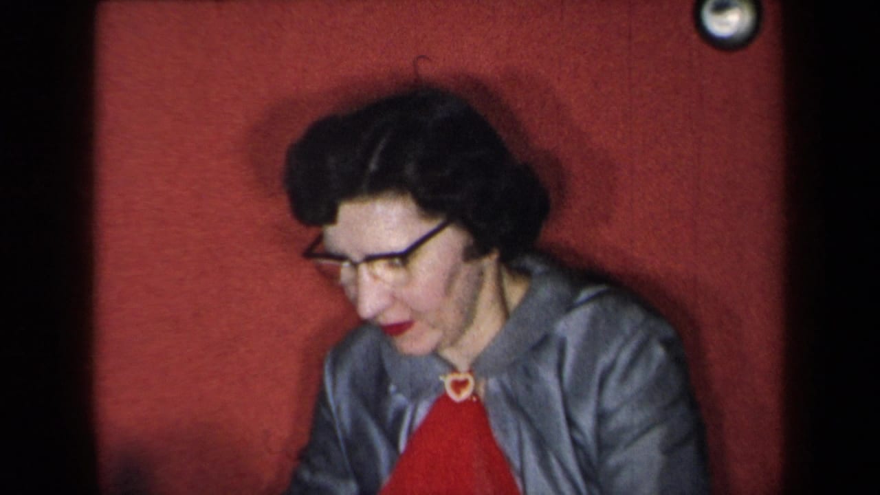 1959 New York home-movie frame of a woman in glasses, gray jacket, and red top seated against a red backdrop.