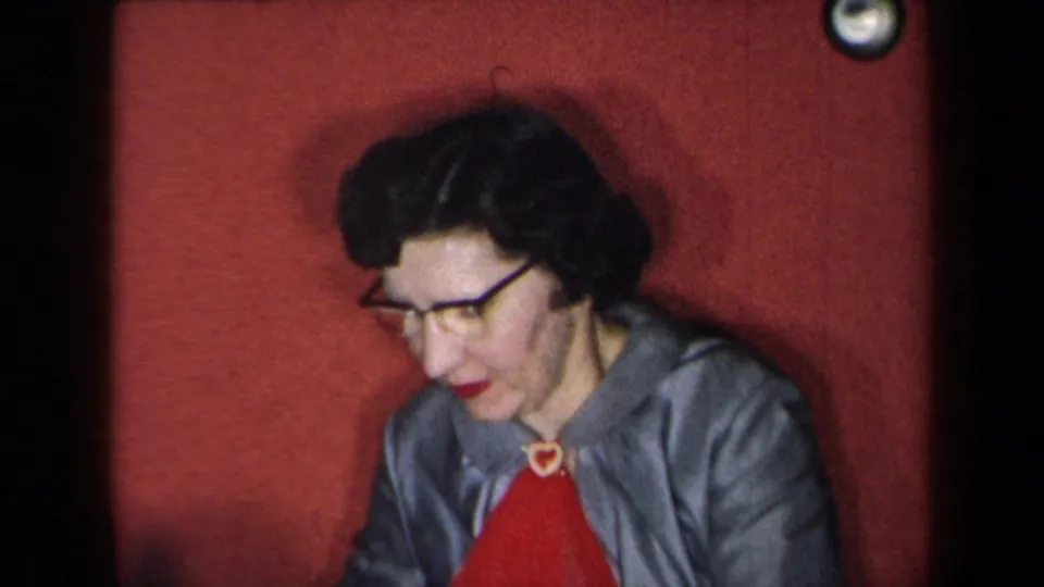 1959 New York home-movie frame of a woman in glasses, gray jacket, and red top seated against a red backdrop.