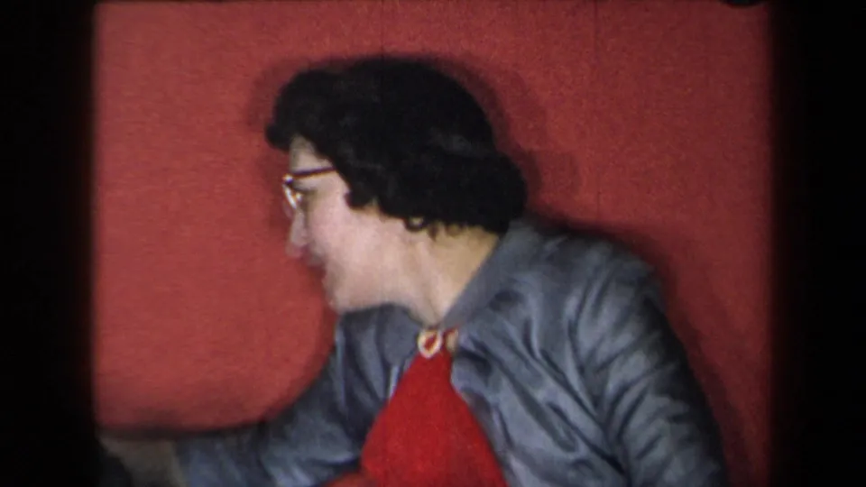 1959 New York 8mm frame showing the woman turned in profile, wearing glasses, a gray jacket, and red top.