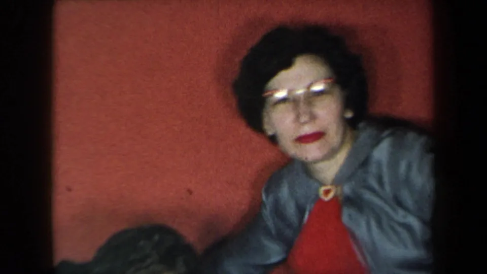 1959 New York home-movie frame of the woman facing the camera with a small smile, seated before a red backdrop.