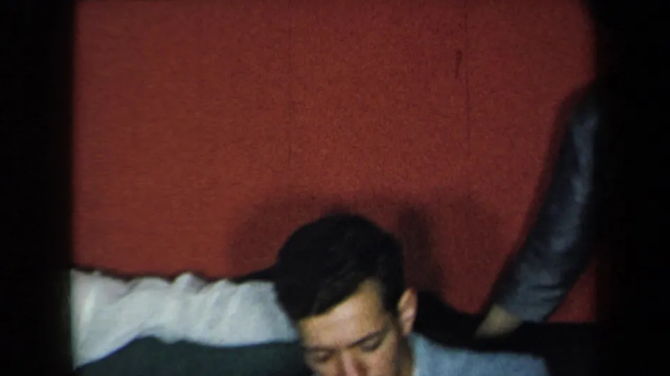 1959 New York home-movie frame showing adults seated or standing near a couch in a red-walled private living room.