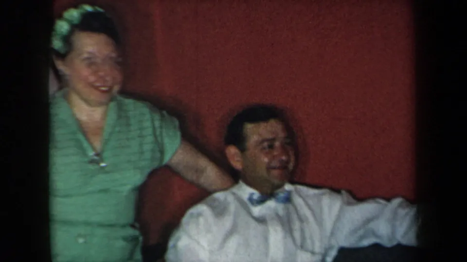 1959 New York interior frame with a smiling woman in green beside a seated man on a couch against a red wall.