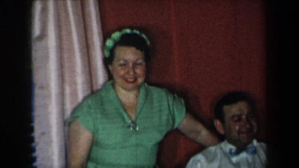 1959 New York home-movie frame showing a woman in a green dress near curtains and a couch in a private living room.