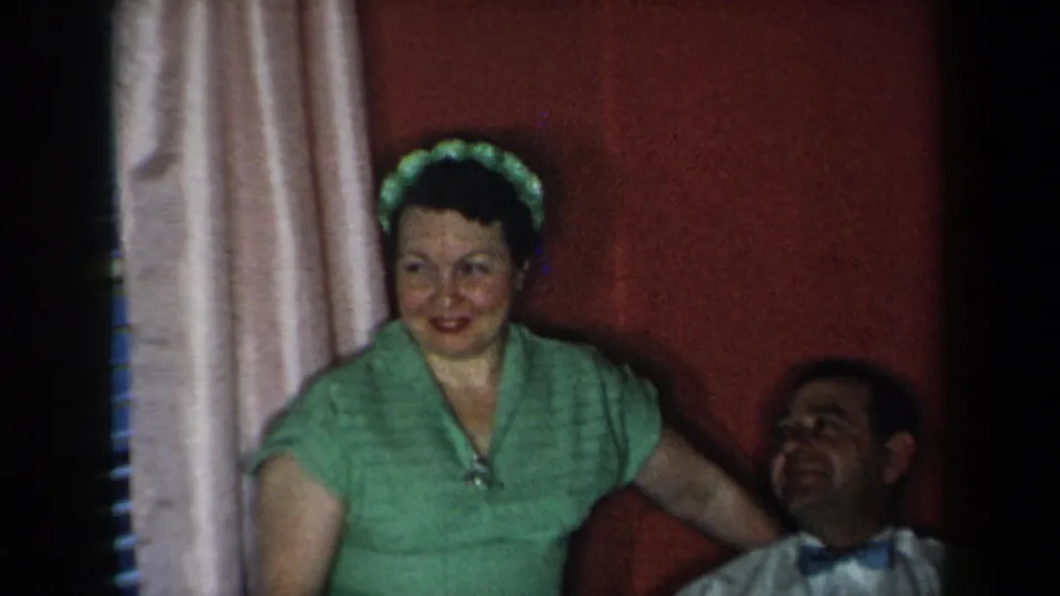 1959 New York living-room frame with a woman in green standing by a seated man against a red wall and pale curtains.