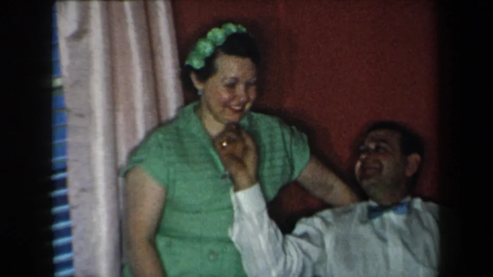 1959 New York home-movie frame showing a woman in green smiling as a seated man laughs near a red wall and curtains.