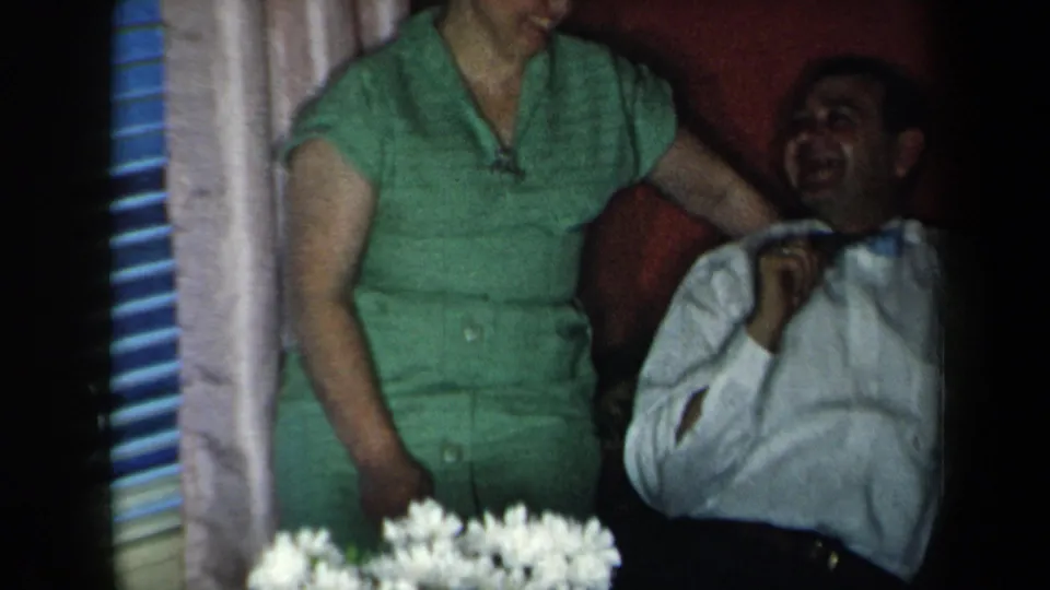 1959 New York home-movie frame with a woman in green standing beside a laughing man, with curtains, blinds, and white flowers visible.
