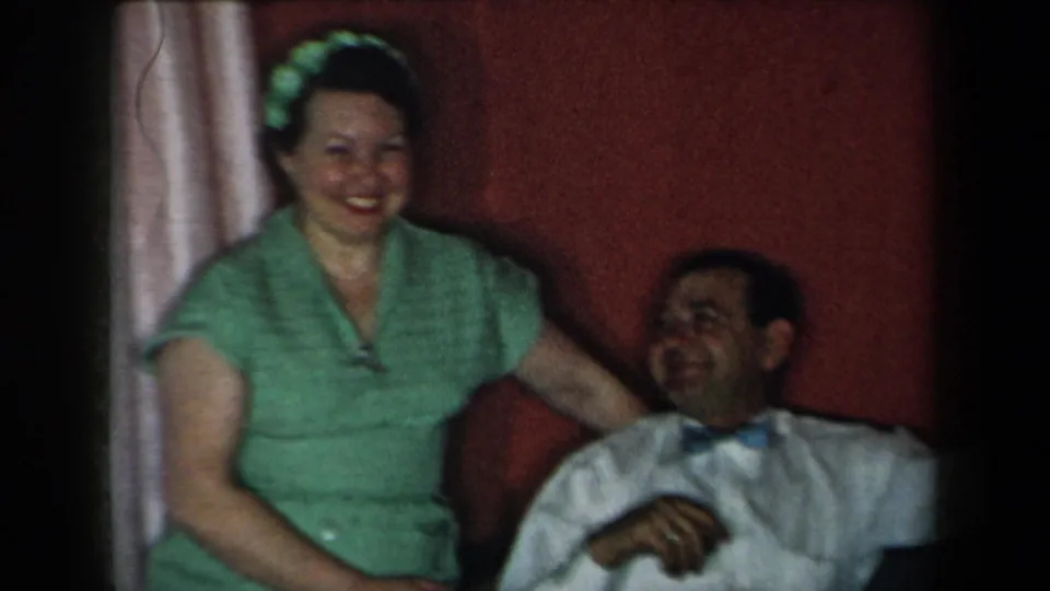 1959 New York home-movie frame of a smiling woman and laughing seated man in a warmly lit domestic room.