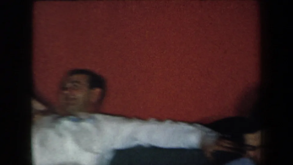 In a 1959 New York home-movie frame, a man in a white shirt leans back near another adult against a red interior wall.