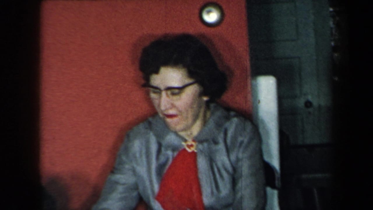 A woman wearing dark glasses and a gray jacket sits by a red wall in a 1959 New York home interior.