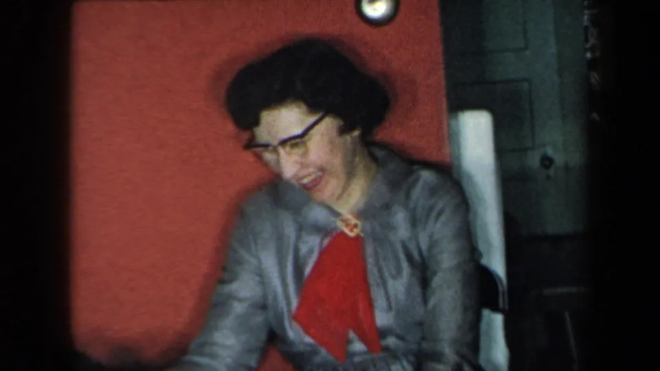 A woman in dark glasses, gray jacket, and red scarf laughs in a 1959 New York home-movie interior.