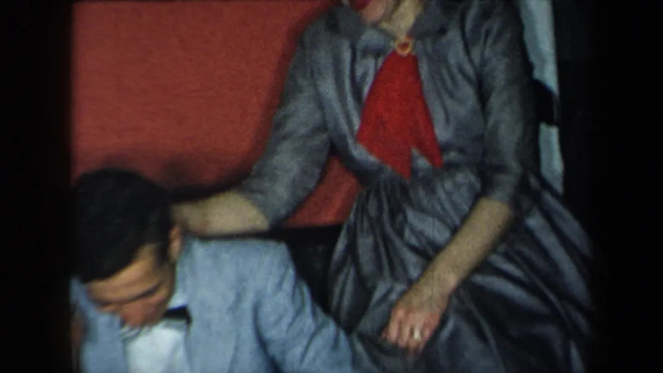 A woman in gray with a red scarf leans beside a seated man in a 1959 New York family home-movie frame.