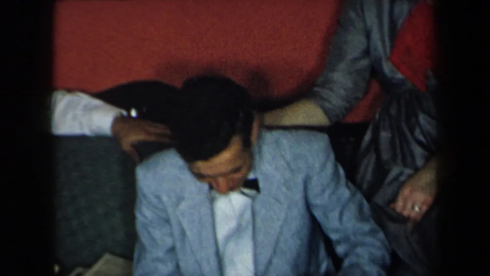 In 1959 New York home-movie footage, a young man in a light blue suit and bow tie sits below red upholstered seating as someone rests a hand near him.