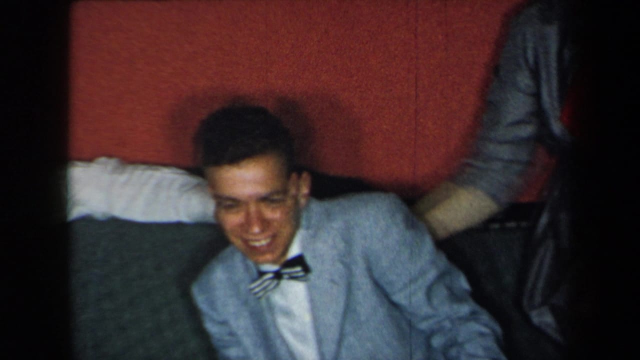 In 1959 New York home-movie footage, the seated young man smiles in a light blue suit and striped bow tie, framed by red upholstery and nearby clothing.