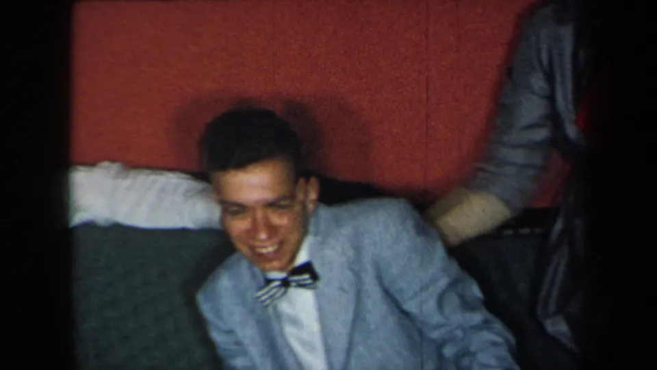 In 1959 New York home-movie footage, the seated young man smiles in a light blue suit and striped bow tie, framed by red upholstery and nearby clothing.