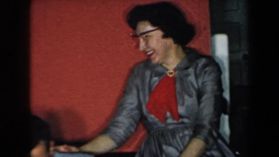 In 1959 New York home-movie footage, a woman wearing glasses, a gray dress, and a red scarf smiles while seated beside the red upholstered backdrop.