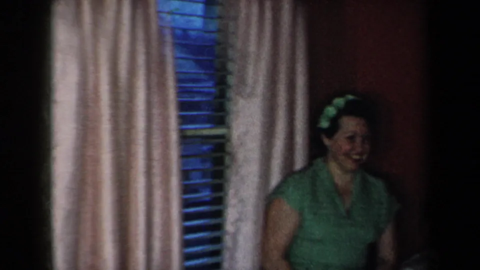 In 1959 New York home-movie footage, a woman in a green dress stands near pale curtains and blue-toned window blinds in a dim interior.