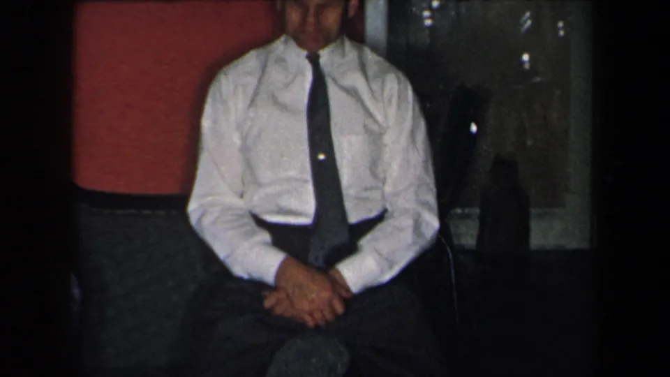In supported 1959 New York home-movie footage, a seated adult man in a white dress shirt and dark tie faces the camera in a private interior.