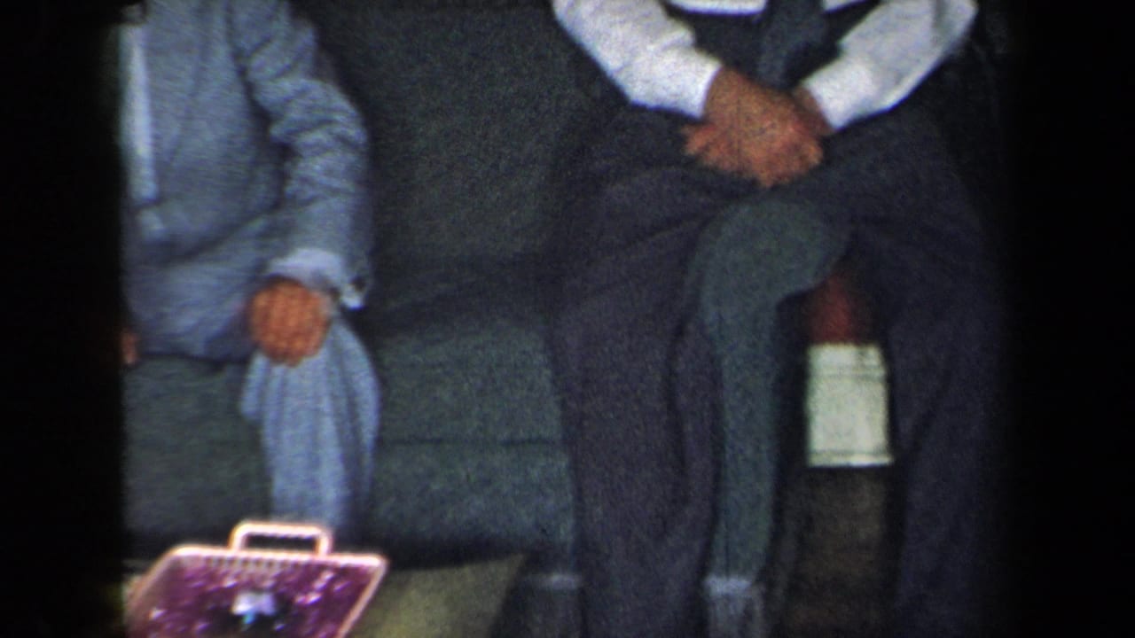 Two formally dressed adults sit on a sofa in the 1959 New York home-movie scene, with a purple basket or container near their feet.