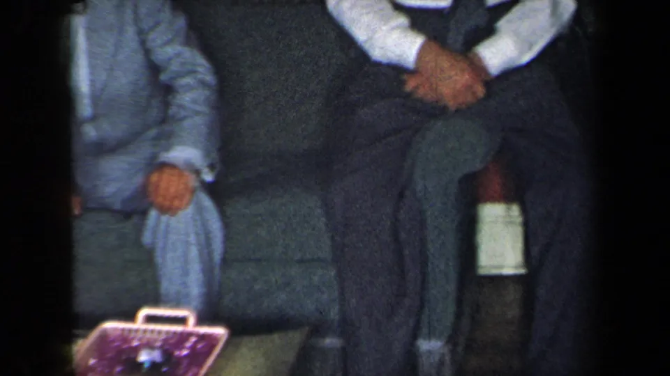 Two formally dressed adults sit on a sofa in the 1959 New York home-movie scene, with a purple basket or container near their feet.