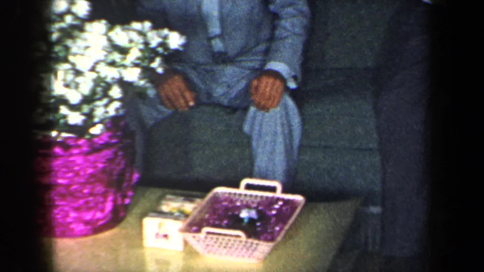 A purple floral container and white flowers dominate the foreground while adults remain partially visible on a sofa in the home-movie interior.
