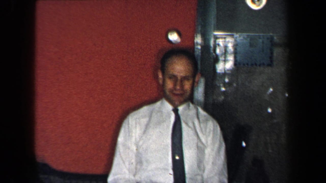 In a 1959 New York home-movie frame, a man stands indoors beside a red wall and dark doorway, facing the camera.