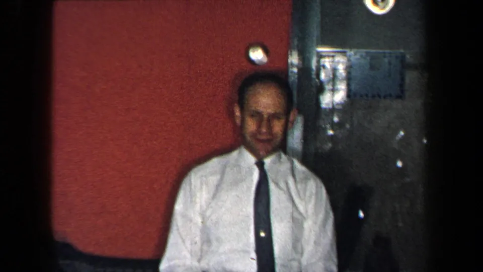 In a 1959 New York home-movie frame, a man stands indoors beside a red wall and dark doorway, facing the camera.